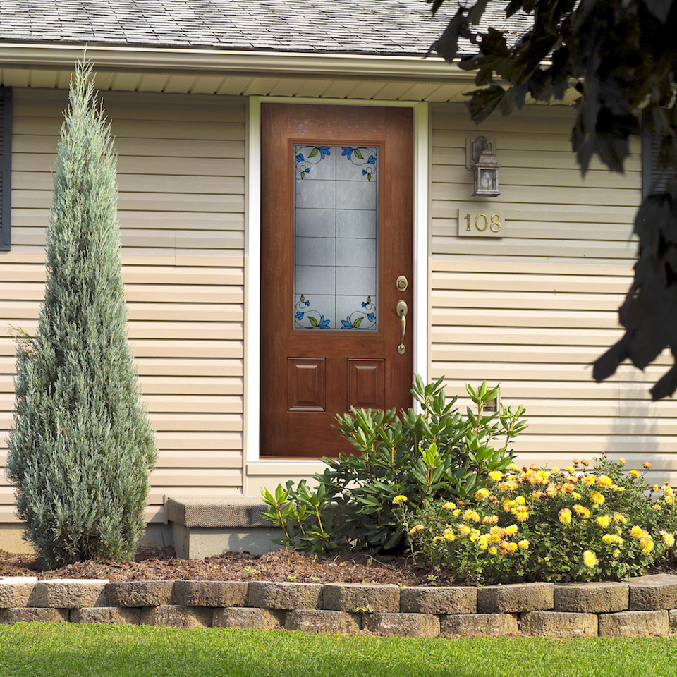 Wood front door with brass hardware and decorative privacy glass on tan house with yellow flowers and bushes in the front.