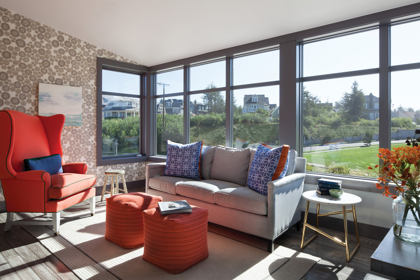 Photo of Andersen E-Series windows in a Dove Gray color. The interior of this living room has medallion print wallpaper, a bright red-orange chair with matching ottomans, a gray couch with blue and red-orange throw pillows. Brennan Enterprises is a premier Andersen Windows and Doors dealer located in North Texas.