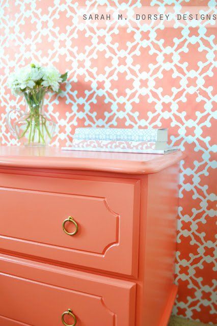 Coral side table with gold ring drawer knobs. The side table is set against a wall featuring coral wallpaper and has flowers as well as books set on top.