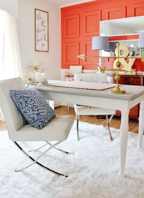 Image of office space with accent wall painted in coral. On the ground is a fluffy white rug with a wooden white desk and two modern white chairs set on top of the rug. Decor includes a navy blue throw pillow on each chair and a colorful pillow on a chair in the background. Other accessories in the image are gold accent pieces as well as a mirror and a sign that says "All the places you'll go".