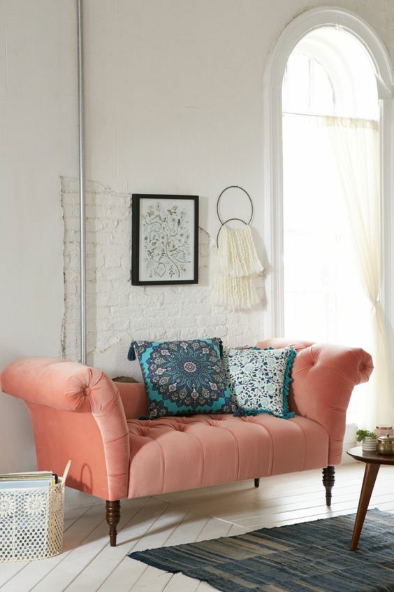 Coral lounge chair from Urban Outfitters. The chair is called the Antoinette Fainting Sofa. The sofa is set against a white wall with wall decor above and two teal blue throw pillows on the sofa. The sofa has wood pegs that match the tone of the wood coffee table in front of the sofa.