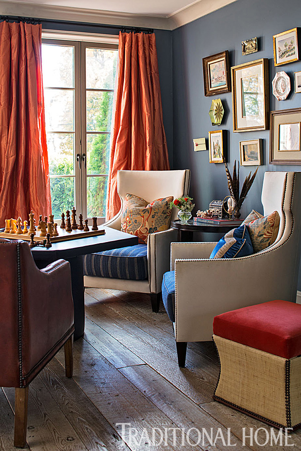 The photo shows an eclectic room with a diverse collection of furniture and decor. The walls in the room are a navy blue, the floors are a weathered oak, the curtains are taffeta fabric in coral.