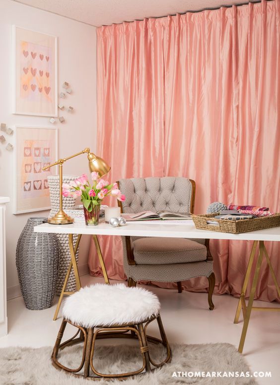 This office space has a youthful and feminine look. It features a coral fabric curtain wall as a backdrop, white walls, a white desk with gold accents, and a gray chair.