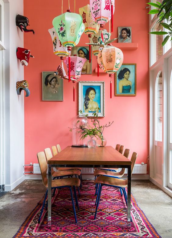 Room with a table that could be a dining room or meeting room. The room has white walls and an accent wall painted in coral. The room features many accessories including a colorful floor rug, hanging lanterns, and wall art.