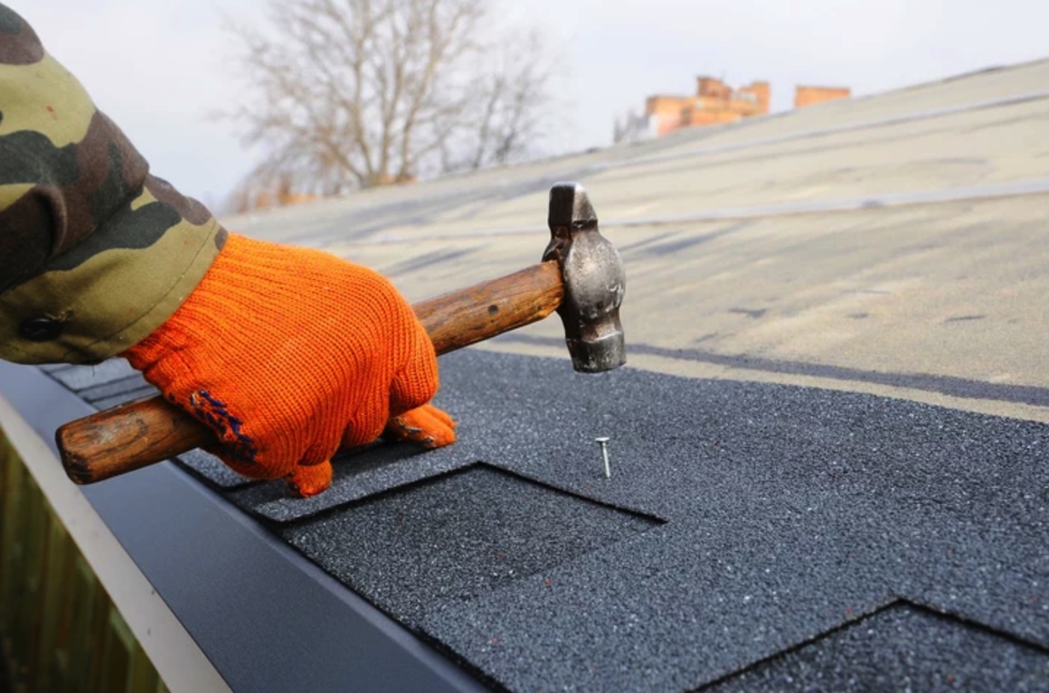 A contractor hammers a nail into an asphalt shingle on a residential roof.