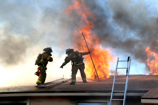 Two firefighters atop a burning residential roof producing large clouds of black smoke.