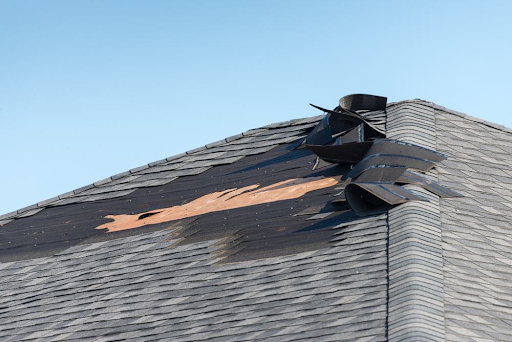 Close up of a roof pitch with several squares of asphalt shingle peeled back from high winds.