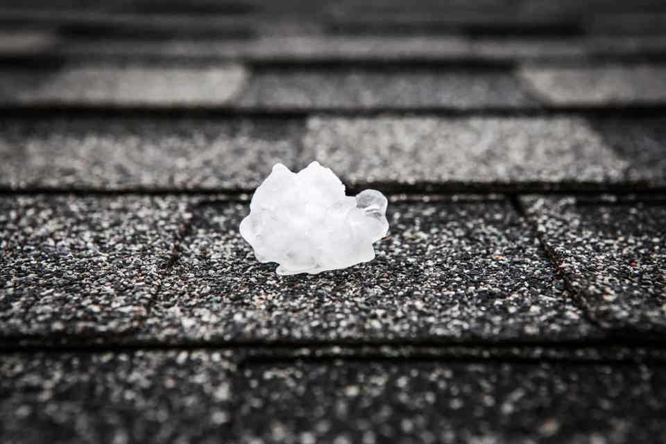 A single piece of white hail atop an asphalt shingle on a roof.